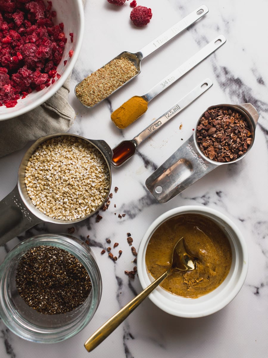 Overhead shot of ingredients needed to make high protein oatmeal, including: raspberries, ground flax seed, cinnamon, vanilla extract, steel cut oats, cacao nibs, chia seeds and almond butter.