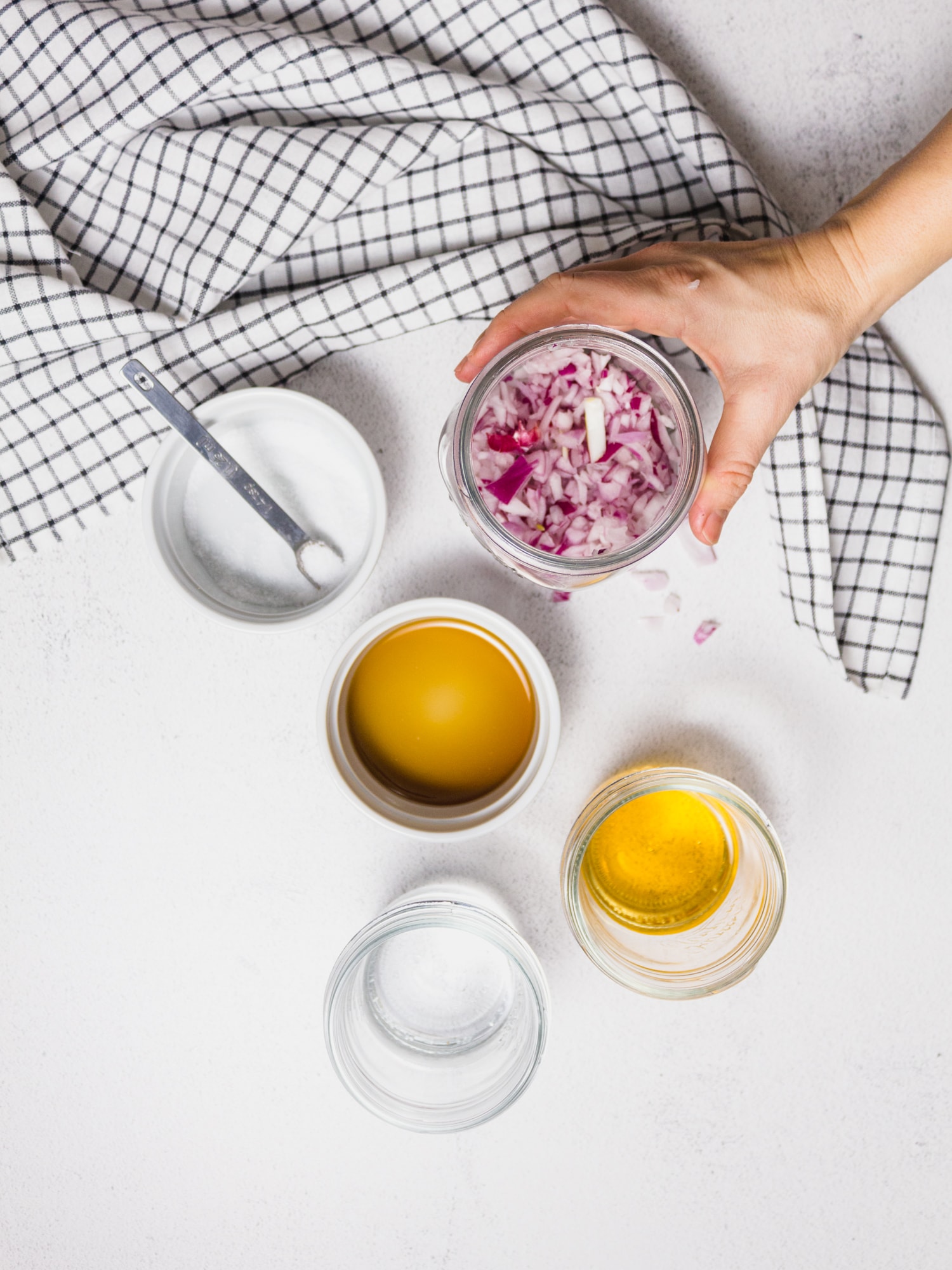 Ingredients needed to make quick pickled red onions. Onions, maple syrup, water, salt and apple cider vinegar are in bowls on a white table. A black and white table cloth is there for decoration.