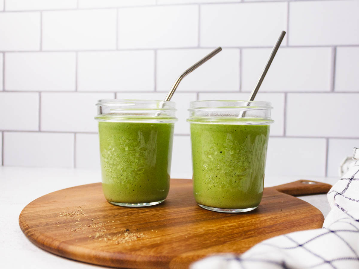 Two mason jars with green vanilla smoothie in them. Both have a metal straw and are placed on a cutting board. 