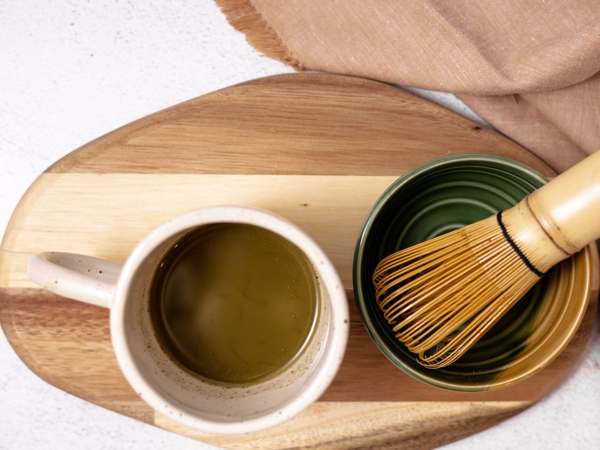 A cup of matcha green tea in a white mug. A bamboo whisk in a green bowl sits next to it. 