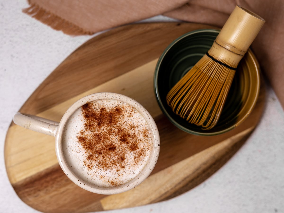 A matcha chai latte in a white mug is on a wooden board. A matcha whisk in a green bowl sits next to it. 