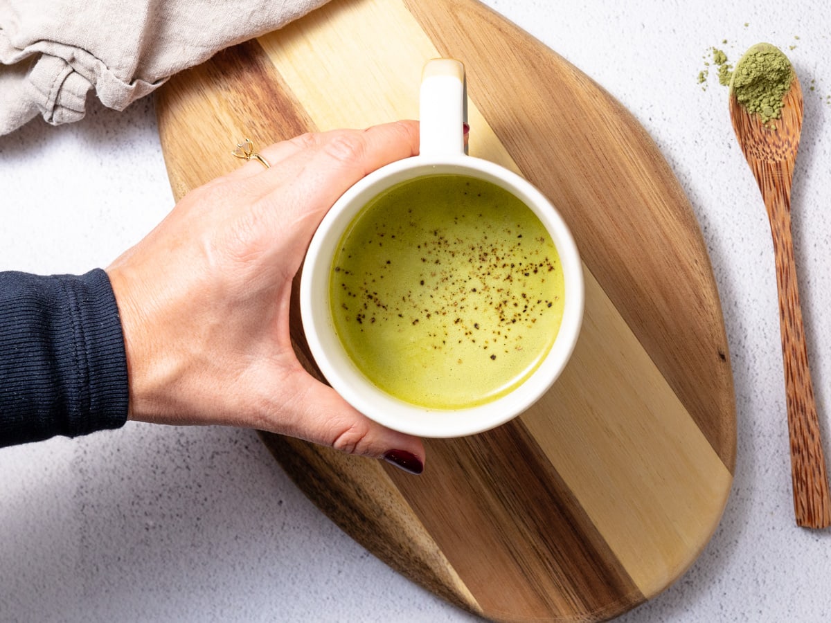 A hand holding a matcha turmeric latte. It's on top of a wooden cutting board.