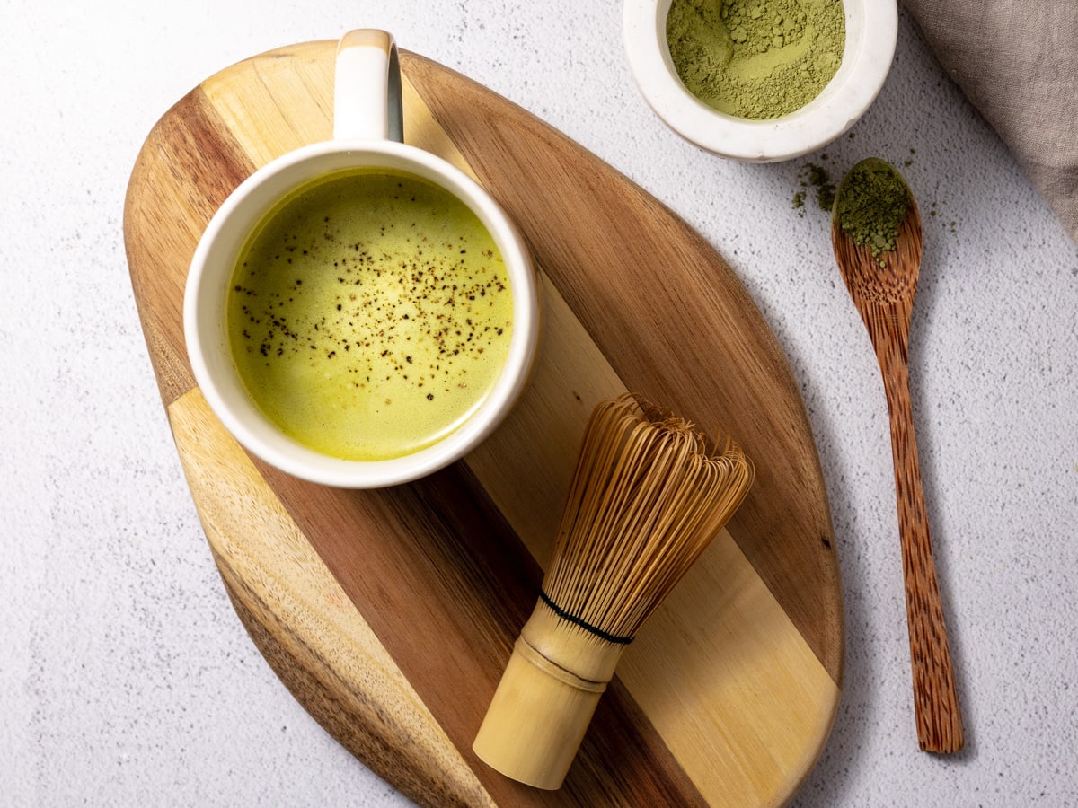 A matcha turmeric latte on a wooden cutting board with a matcha whisk and spoonful of matcha powder beside it.