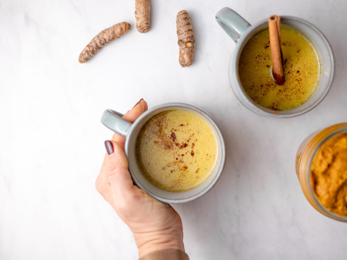 A hand holding a mug of golden milk. Some tumeric root is on the white countertop as well.