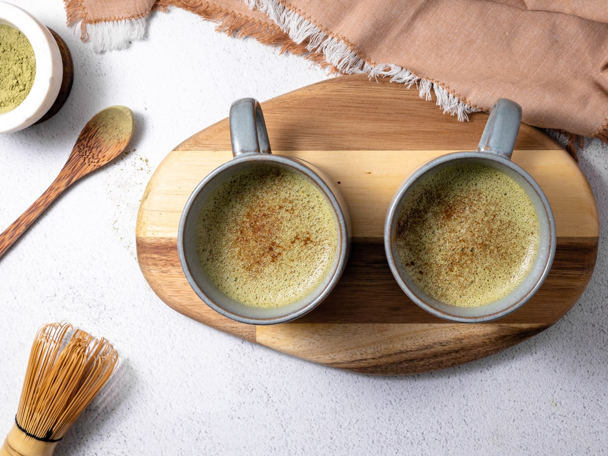 2 mugs filled with vanilla matcha latte are sitting on a wooden cutting board. A matcha whisk and spoonful of matcha powder sit near by. 