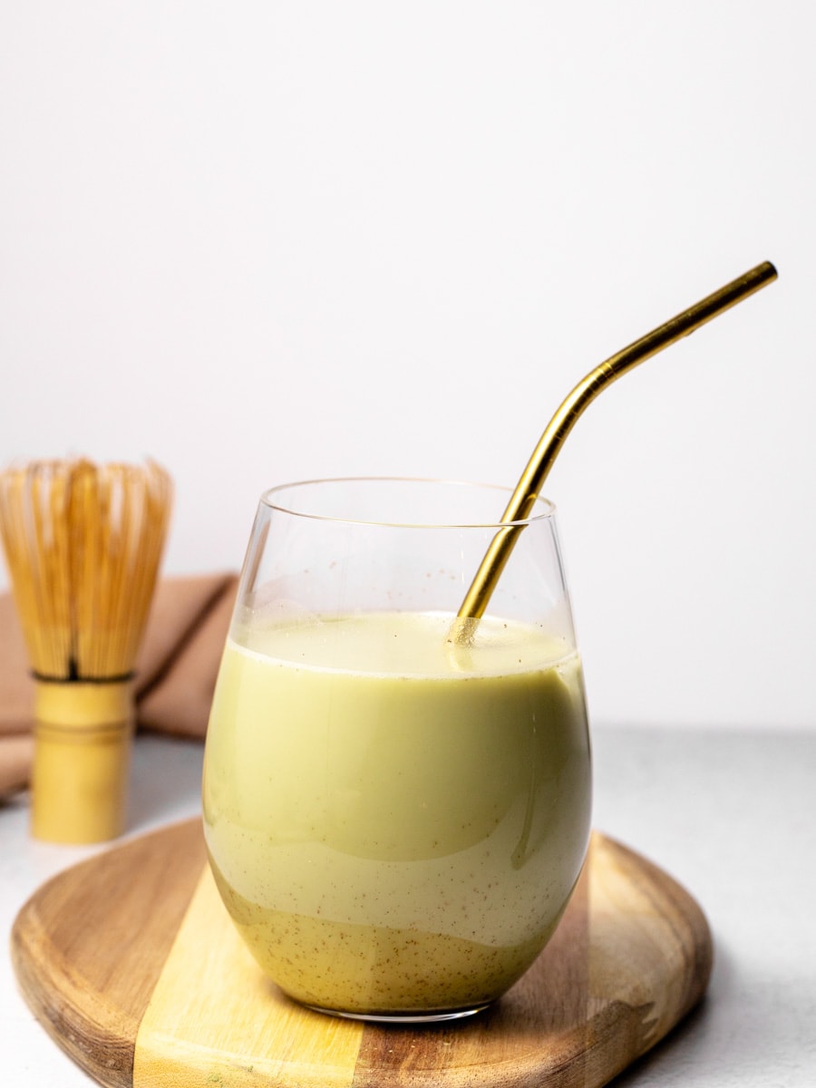 An iced matcha vanilla latte with a gold straw sits on a wooden cutting board. 