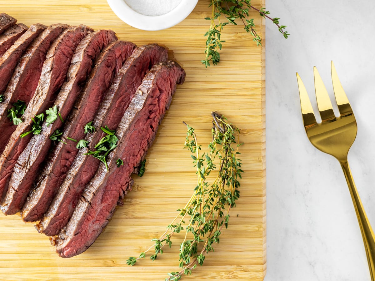 A sous vide flank steak resting on a wooden cutting board. A gold fork sits by its side. 
