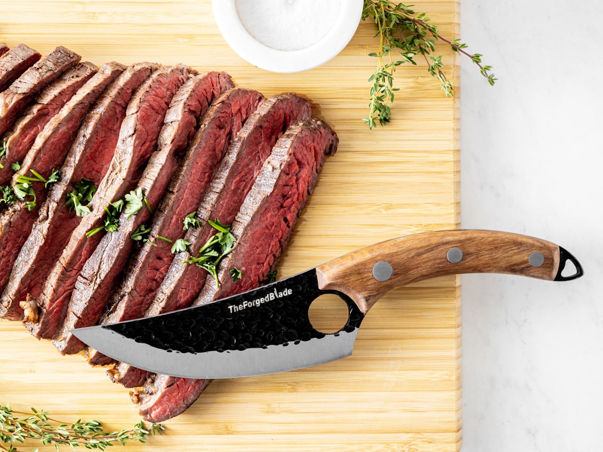 The forged blade resting on top of a flank steak on a wooden cutting board. 