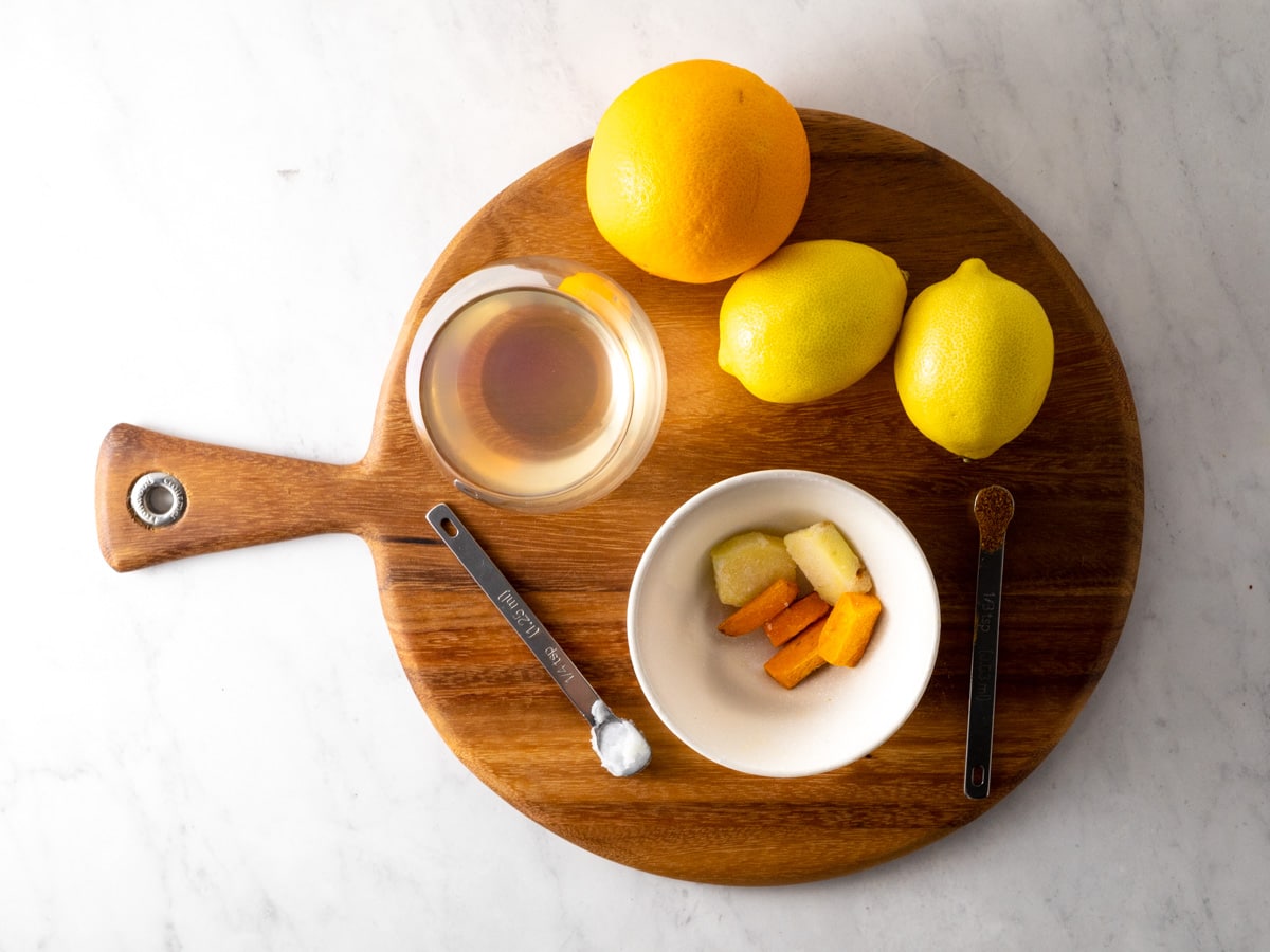 Ingredients needed to make a turmeric juice shot. Lemons, orange, coconut water, turmeric, ginger, coconut oil and cayenne pepper. All sitting on a wooden cutting board. 
