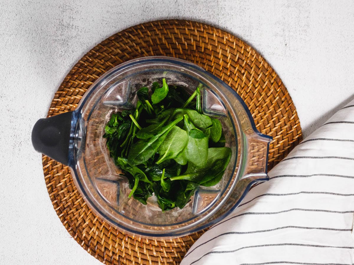 A vitamix blender with green spinach in it. On a wooden placemat. 