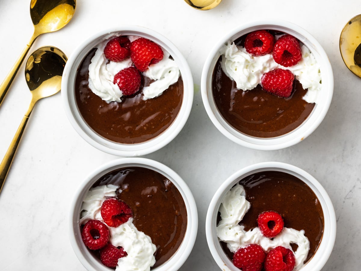 4 ramekin containers with chocolate mousse in them. Gold spoons lay on the countertop next to them.