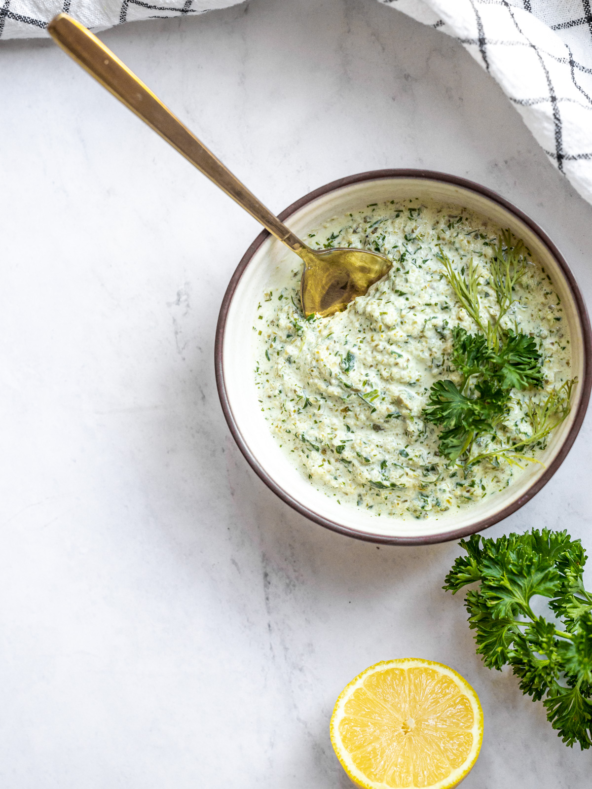 A bowl of keto tartar sauce on a white countertop with a gold spoon in it. A striped black and white cloth is placed nearby. 