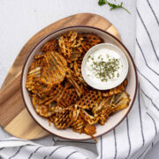 A bowl of waffle fries on a wooden cutting board with a striped dish cloth nearby.