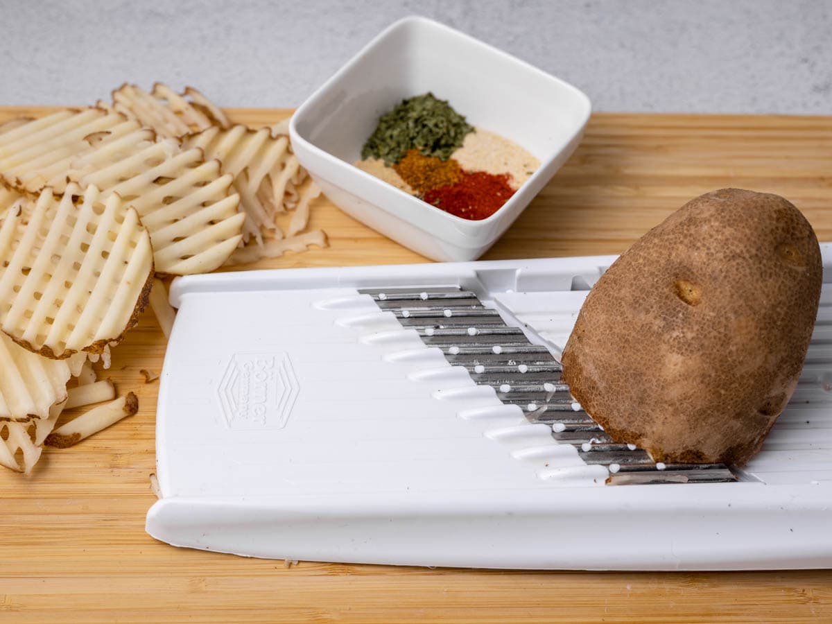 A potato being sliced on a wave waffle cutter on top of a wooden cutting board.