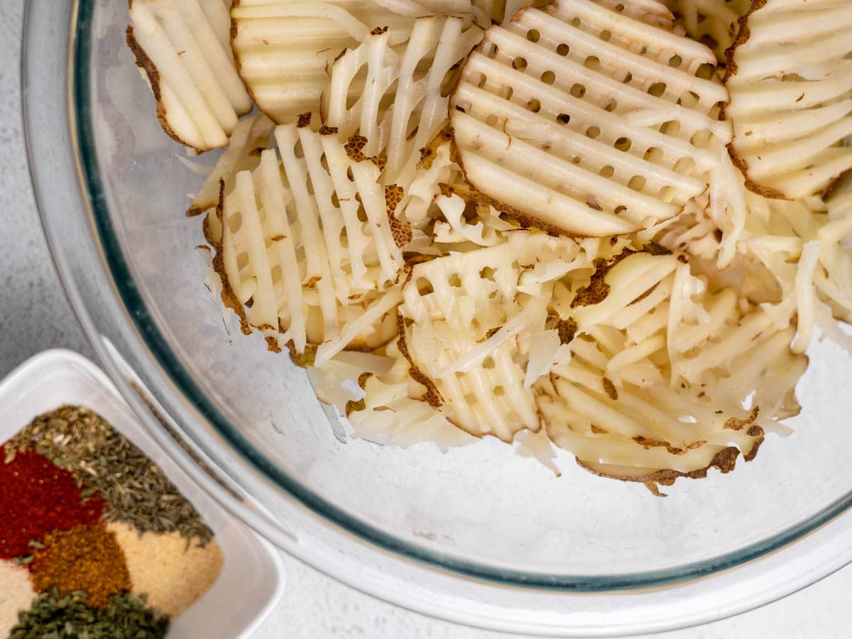 A bowl of uncooked waffle fries on a white countertop with a seasoning mix to the left.