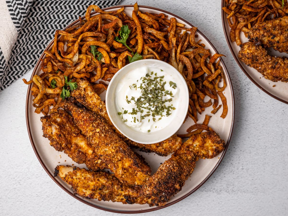 A plate of chicken fries and curly fries with a white dipping sauce. Sitting on a white countertop.