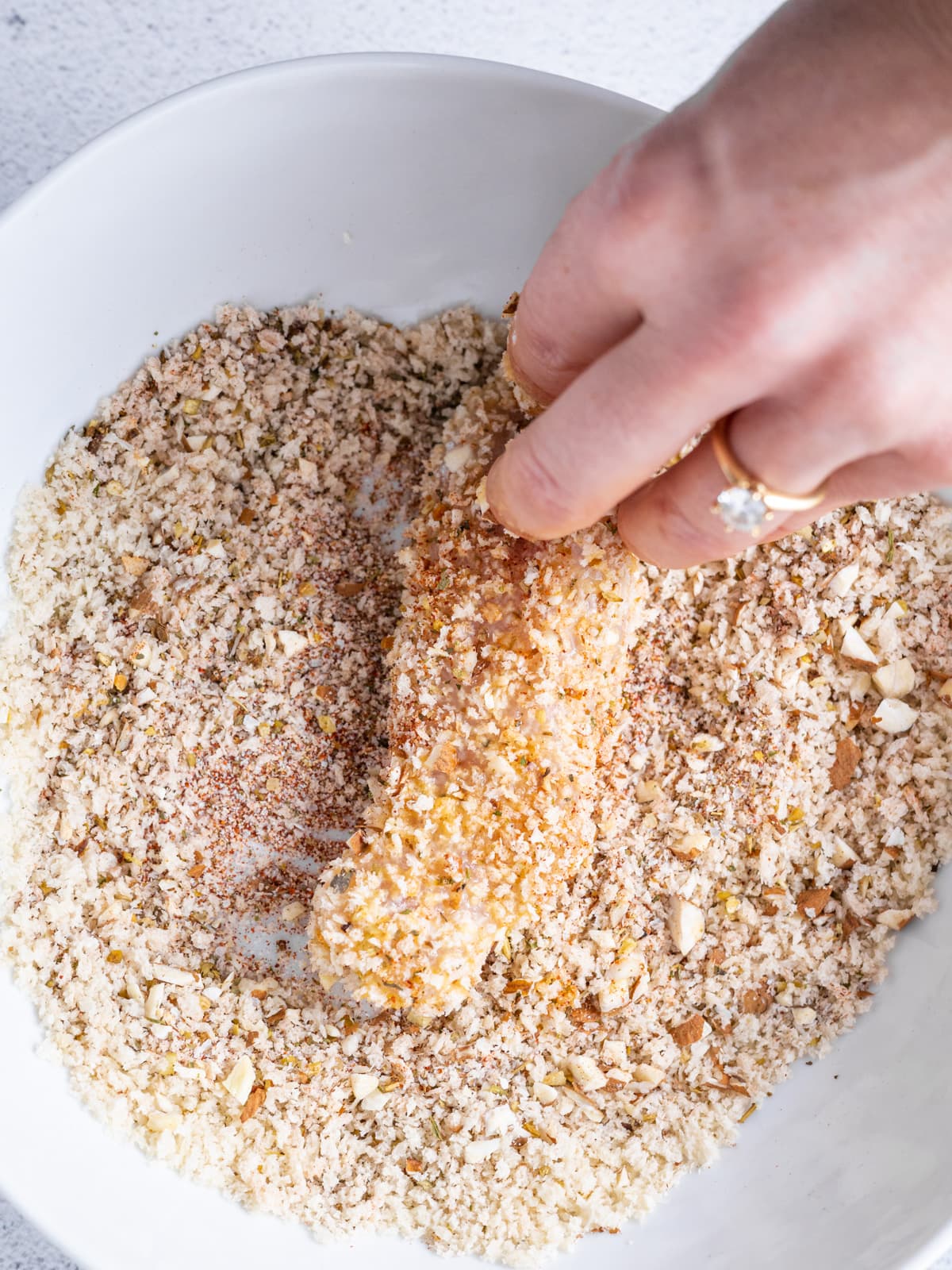 A chicken breast being dipped in breading in a white bowl.