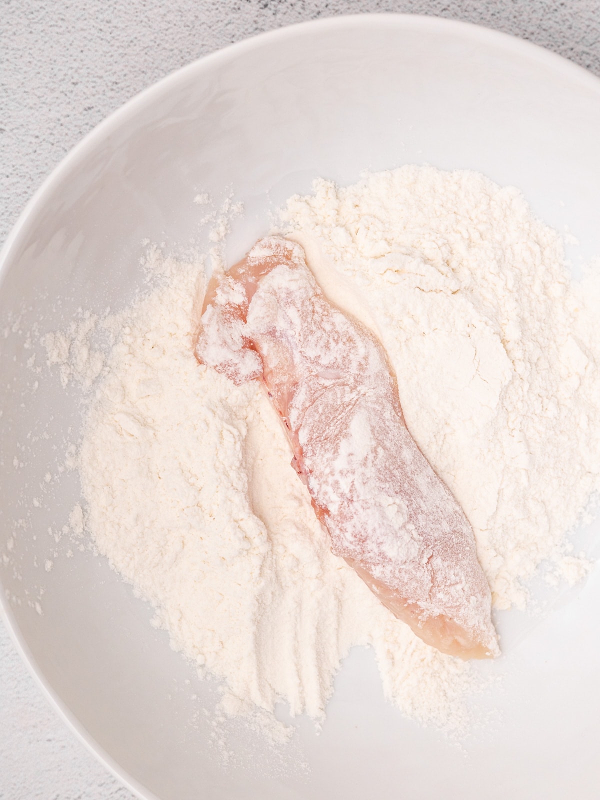 A chicken breast in flour in a white bowl.