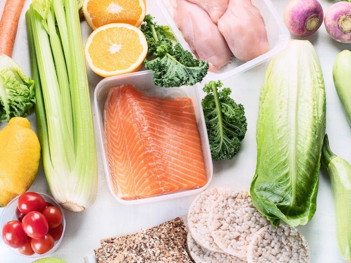 Overhead shot of a variety of food on a white countertop, including celery, lemons, salmon, raw poultry and lettuce. 