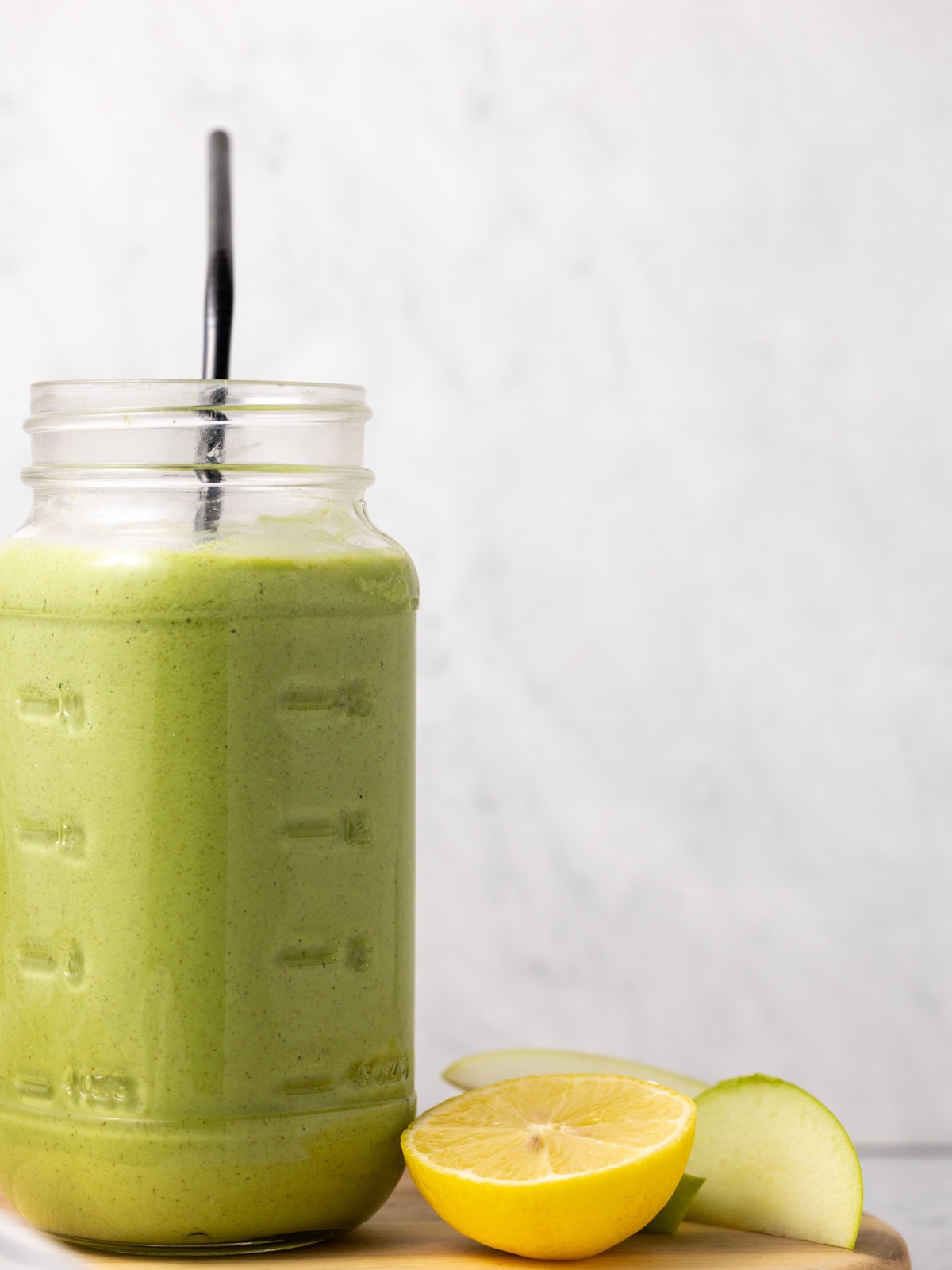 A green smoothie in a large mason jar. A lemon and apple slices sit beside it. 