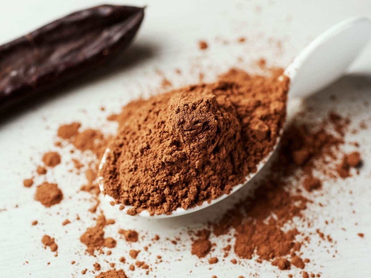 A spoonful of carob powder on a white countertop. A carob bean sits nearby.