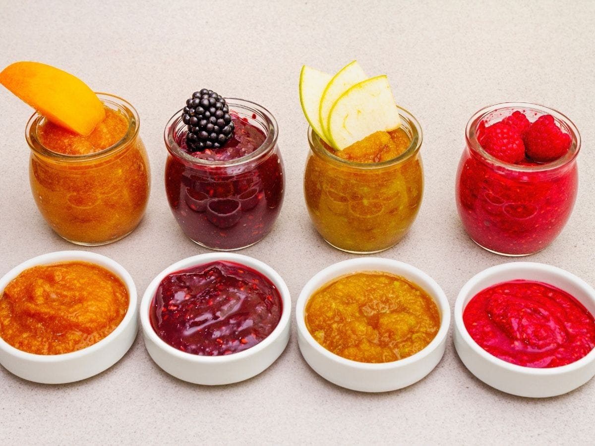 Four jars of fruit puree. Four small white bowls of fruit puree sit in front of them.
