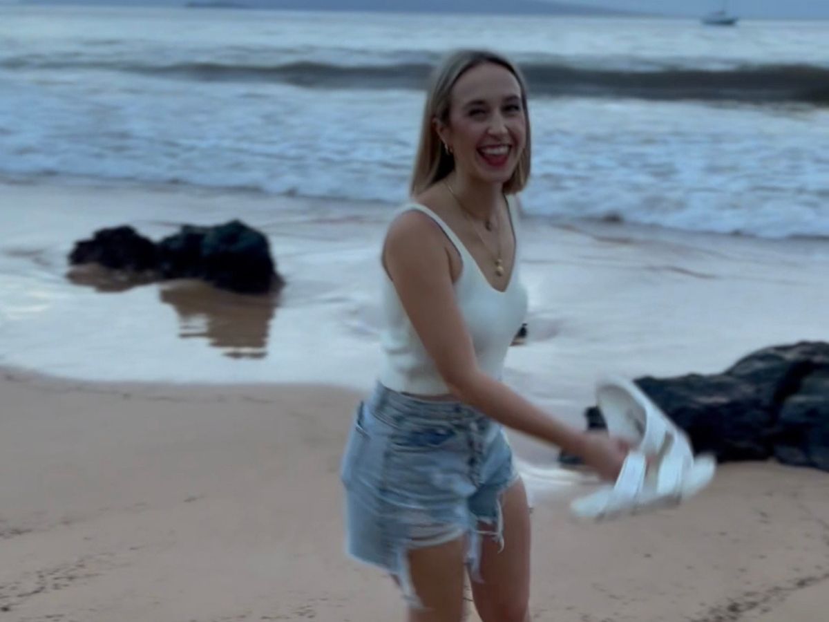 A girl at the beach holding her sandals. 