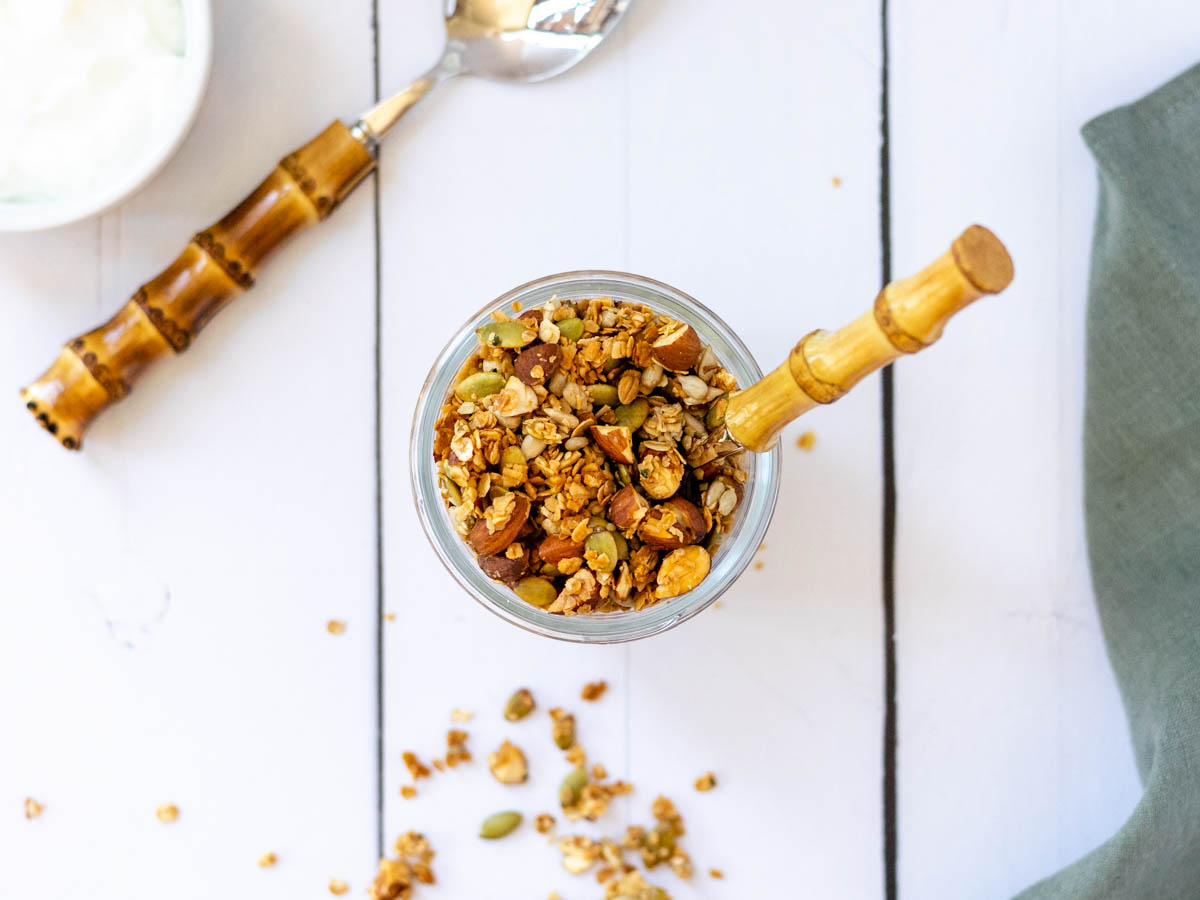 A glass jar with granola in it, with a bamboo spoon coming out. On a white wooden tabletop.