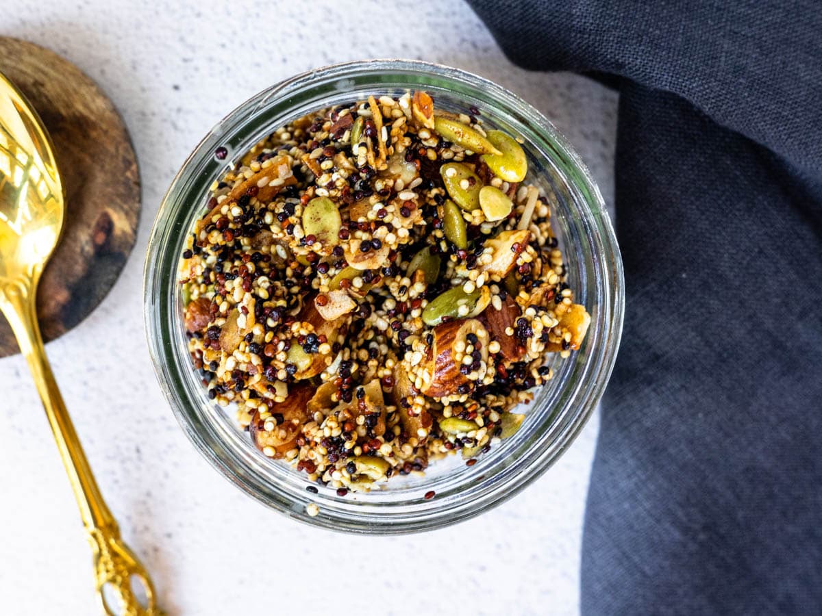 Almond vanilla granola in a glass mason jar with a gold spoon and dark blue linen cloth nearby.