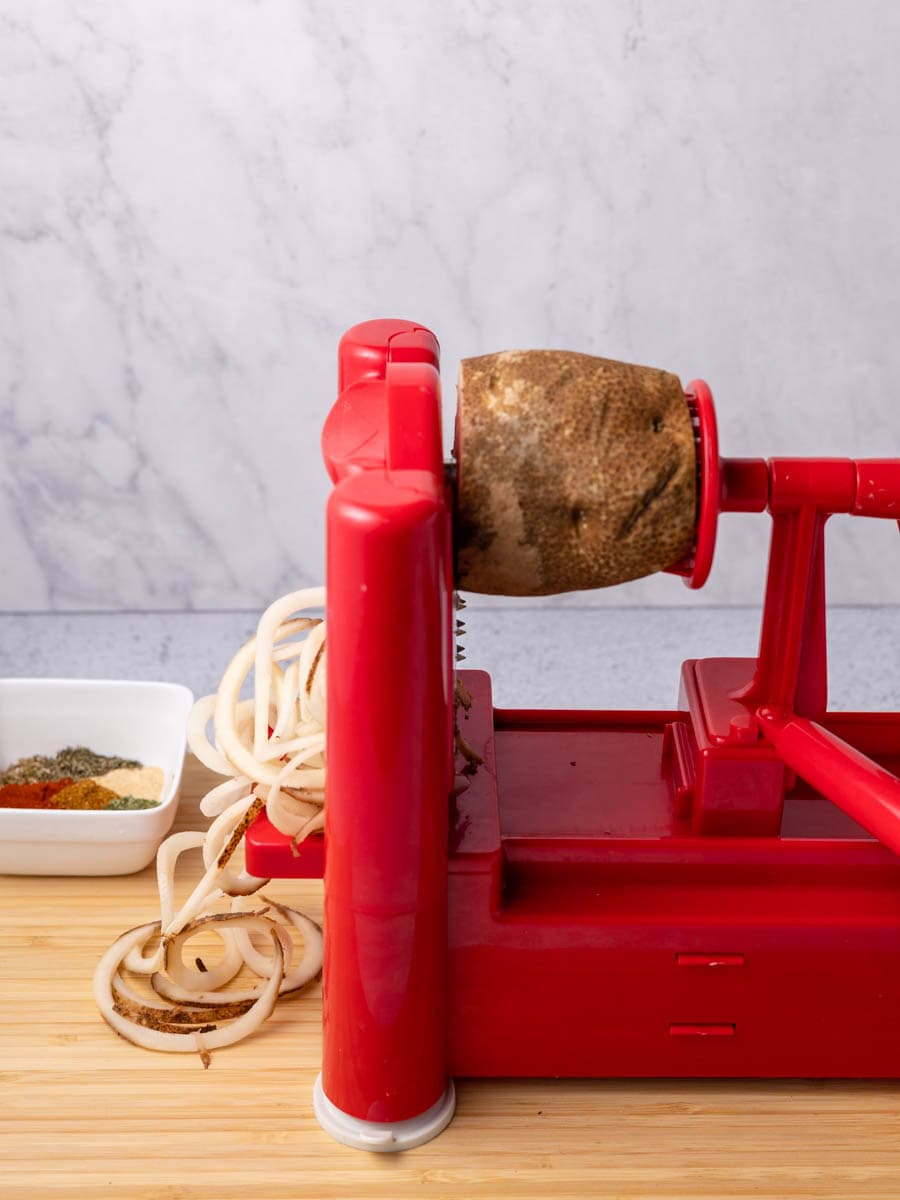 Potato on a red spiralizer, being spiralized onto a wooden cutting board. 