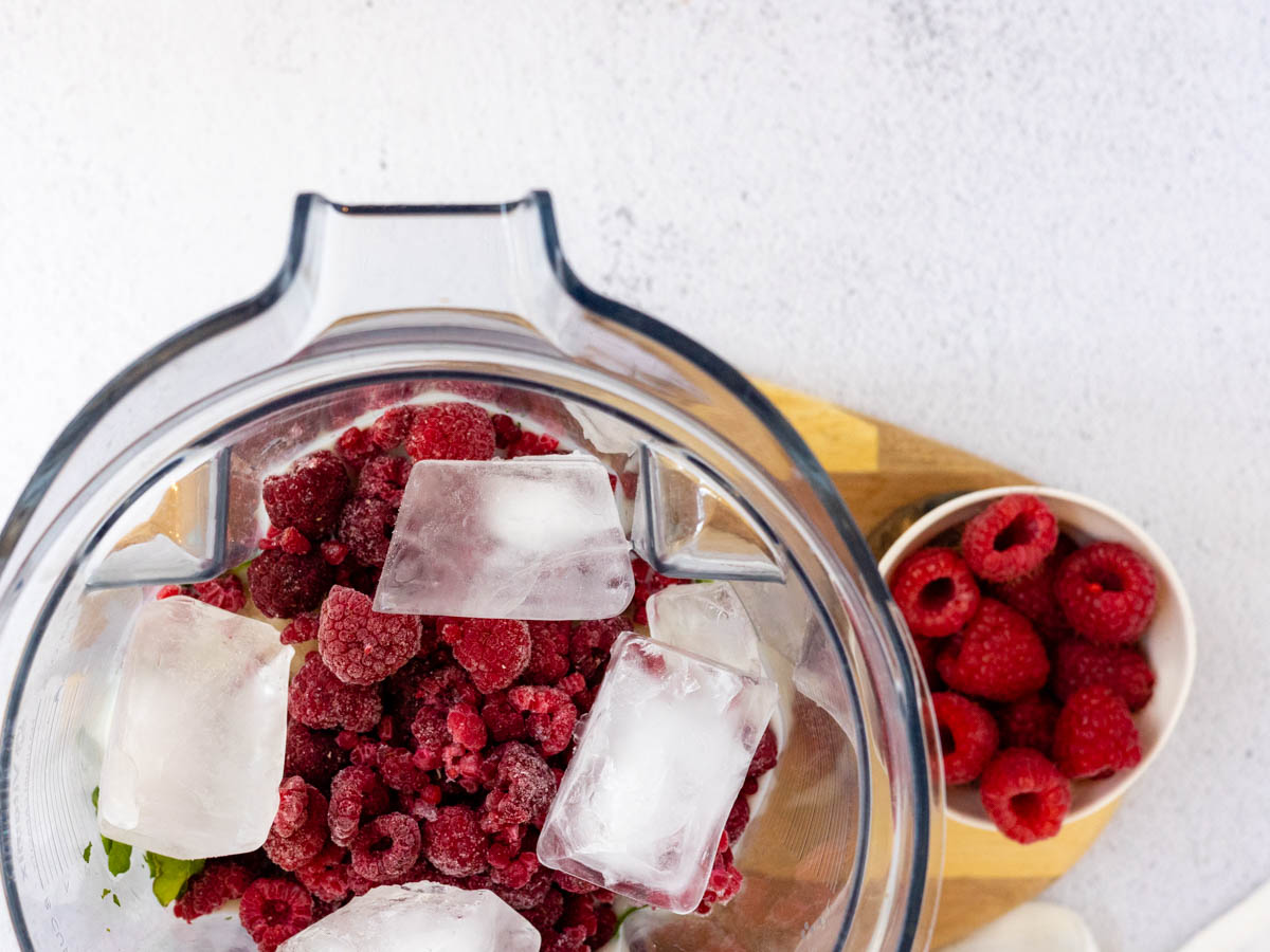 frozen raspberries and ice cubes in a blender on a wooden cutting board.