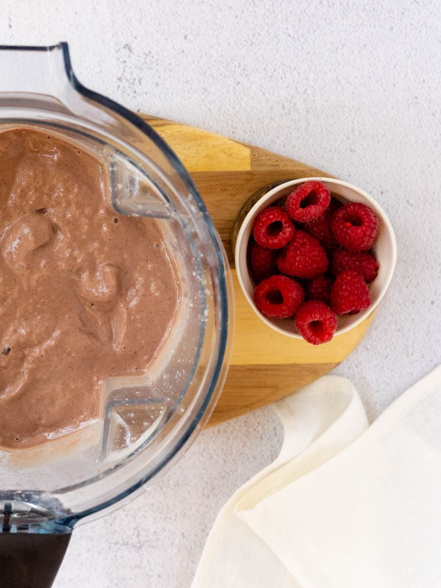 An almond milk raspberry smoothie blended in a vitamix blender. A cup of raspberries sit nearby. 