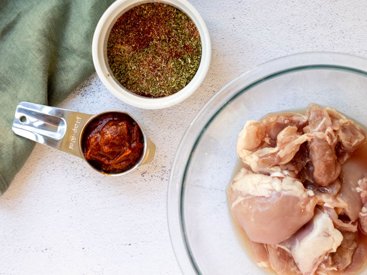 BBQ sauce, spice rub, and raw chicken thighs on a white countertop with a green linen napkin nearby.