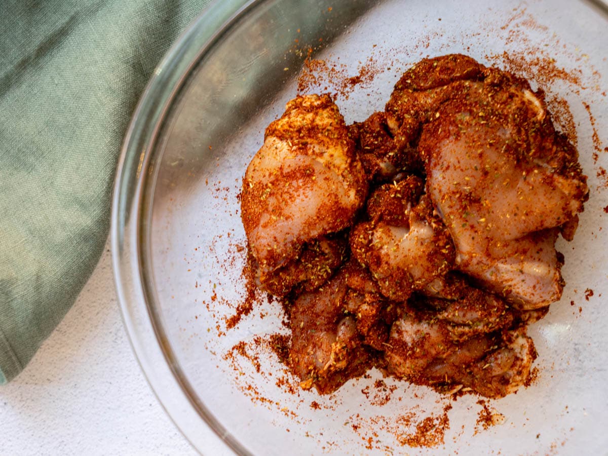 Chicken thighs covered in a spice rub in a glass bowl.