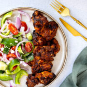 Air fryer bbq chicken thighs on a ceramic plate with a greek salad. Gold cutlery lies nearby.