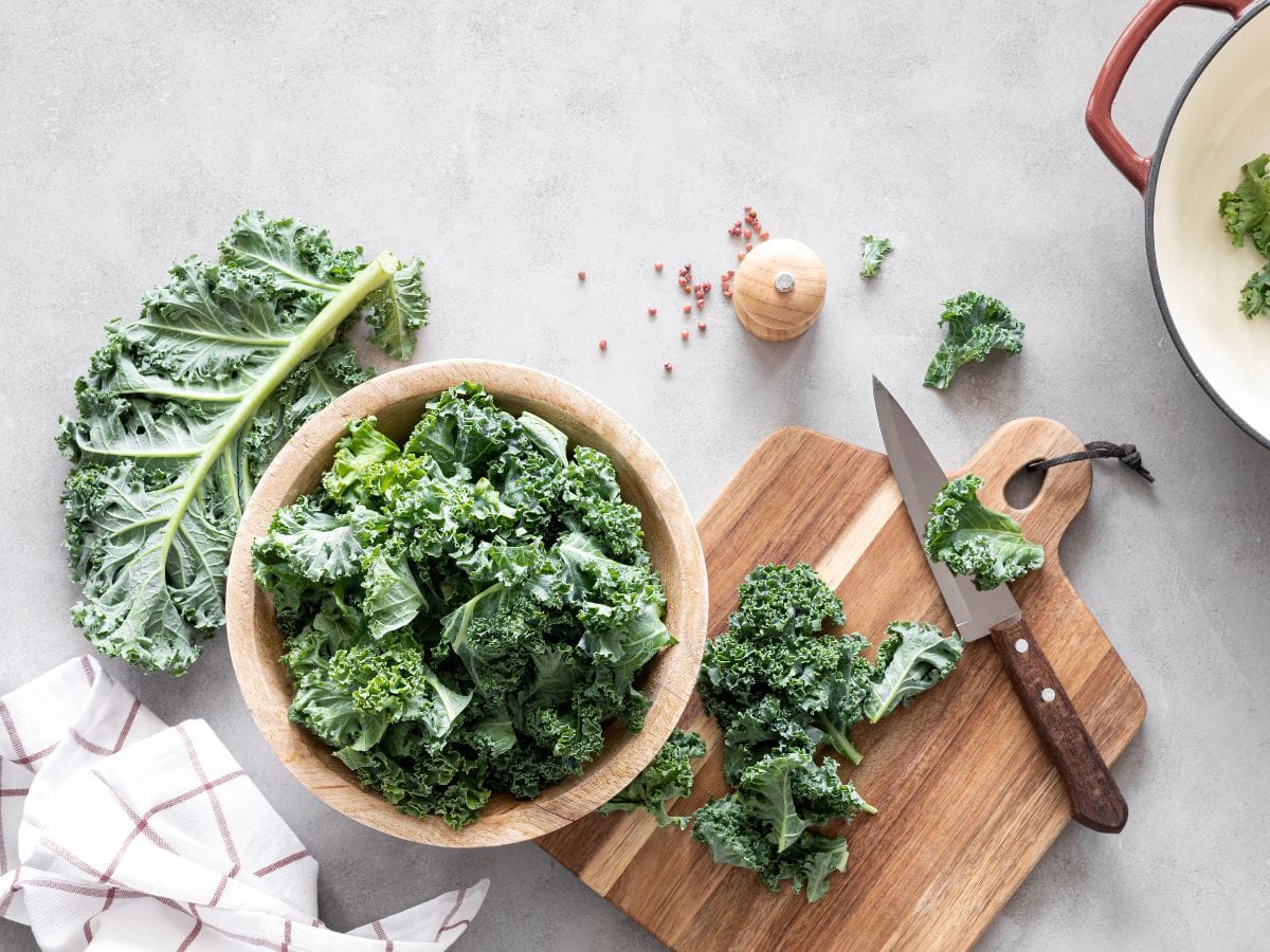 Kale in a wooden bowl, with a piece of kale being cut on a wooden cutting board.