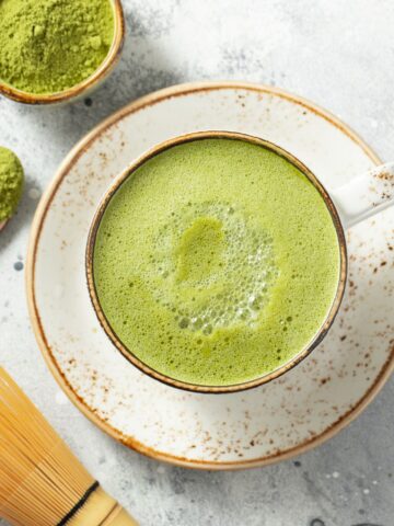 A bowl of matcha powder, a matcha drink, and a bamboo whisk on a white countertop.