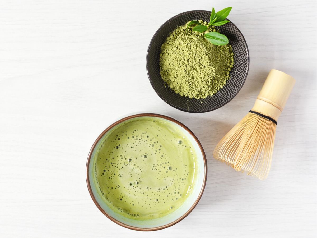 A bowl of matcha powder, whisked matcha, and a bamboo whisk on a white countertop.