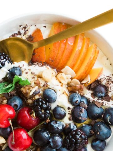 A yogurt bowl on a white counter with blueberries, peaches, cherries and mixed nuts.