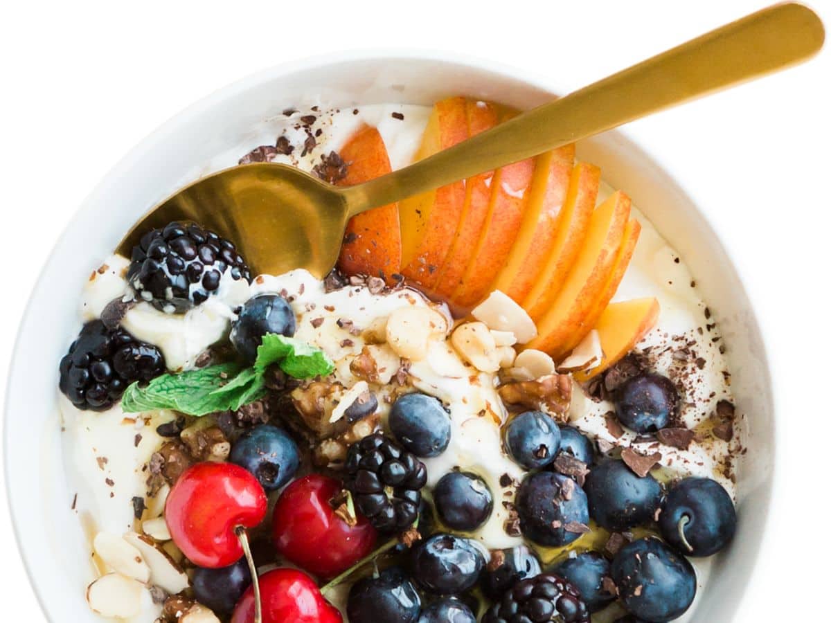 A yogurt bowl on a white counter with blueberries, peaches, cherries and mixed nuts.