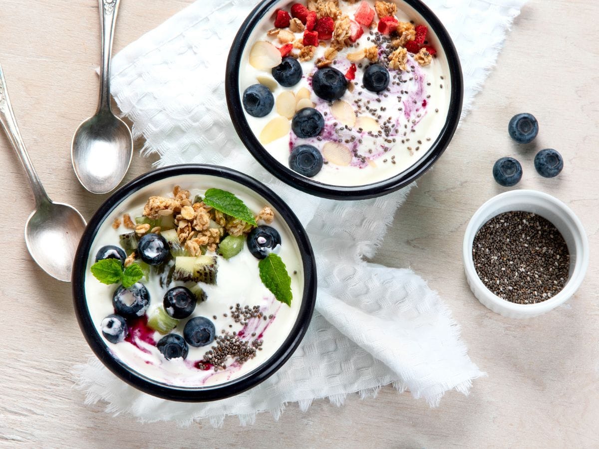 Two yogurt bowls on white counter with a variety of ingredients in them. Two silver spoons lay beside.