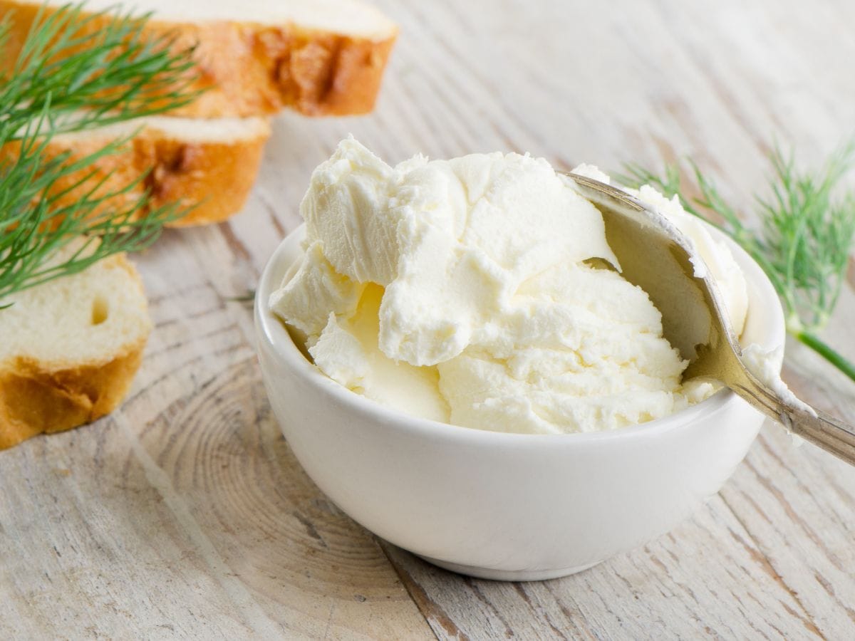A bowl of cream cheese on a wooden countertop with slices of bread nearby.,