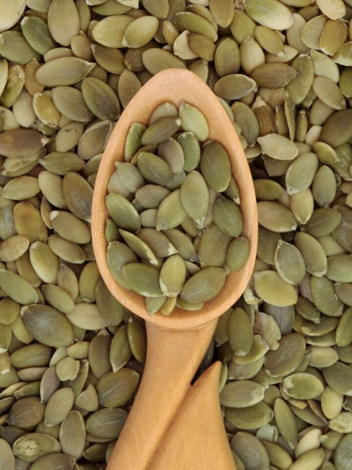 A wooden spoon with pumpkin seeds in it surrounded by more pumpkin seeds.